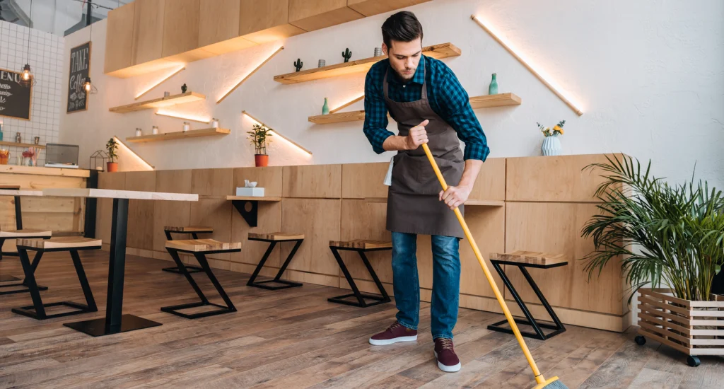 young-worker-in-apron-cleaning-floor-with-sweep-in-2022-12-16-15-39-28-utc